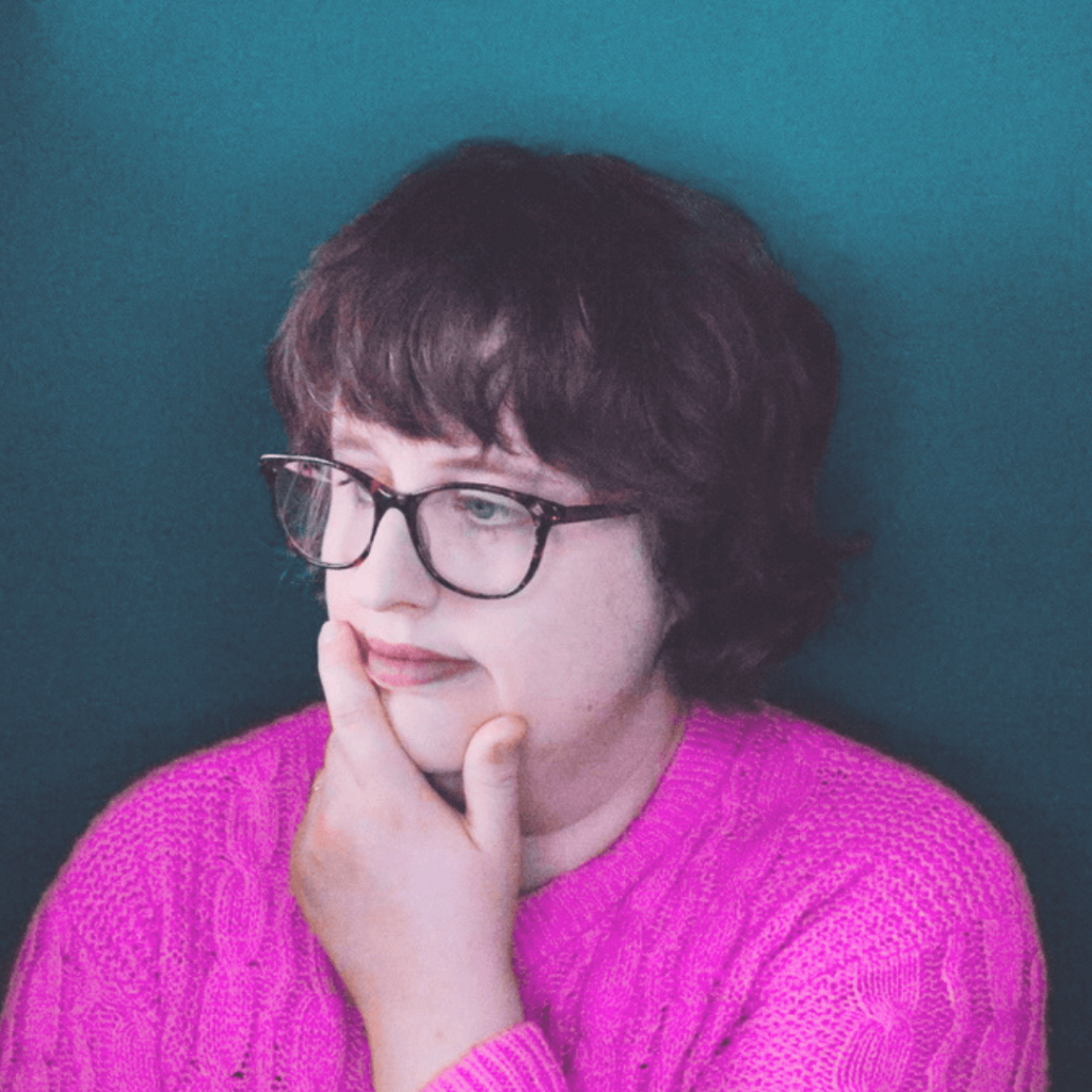 A person with chin-length brown hair and glasses, wearing a vibrant pink jumper, is looking thoughtfully to their left with their chin resting on their hand. They are in a room with a teal-coloured wall